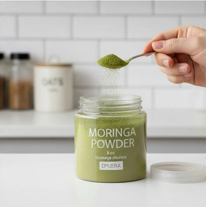 Jar of Druera moringa powder with a spoonful being added, on a kitchen counter.